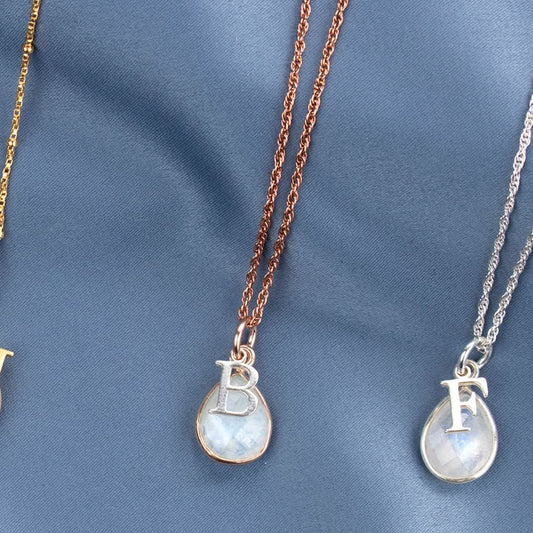 two moonstone charm necklaces in silver and rose gold  with silver initial charms photographed on blue satin fabric