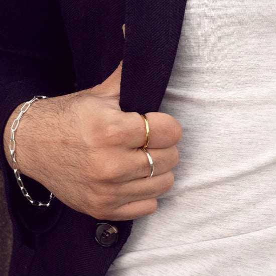 A man wearing a gold and silver ring with his hand on his blazer and a paperclip bangle round his wrist 