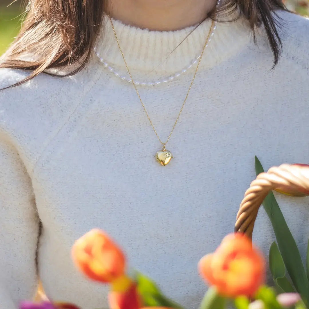 A person wearing a gold heart-shaped locket necklace over a cream-colored sweater, with a basket of tulips in the foreground.