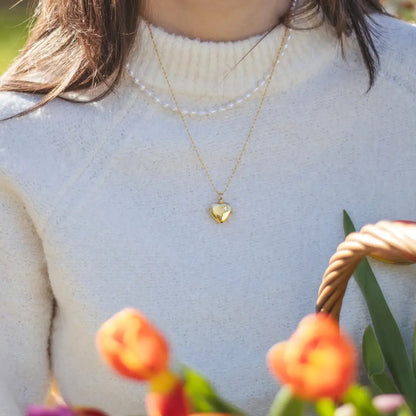 A person wearing a gold heart-shaped locket necklace over a cream-colored sweater, with a basket of tulips in the foreground.
