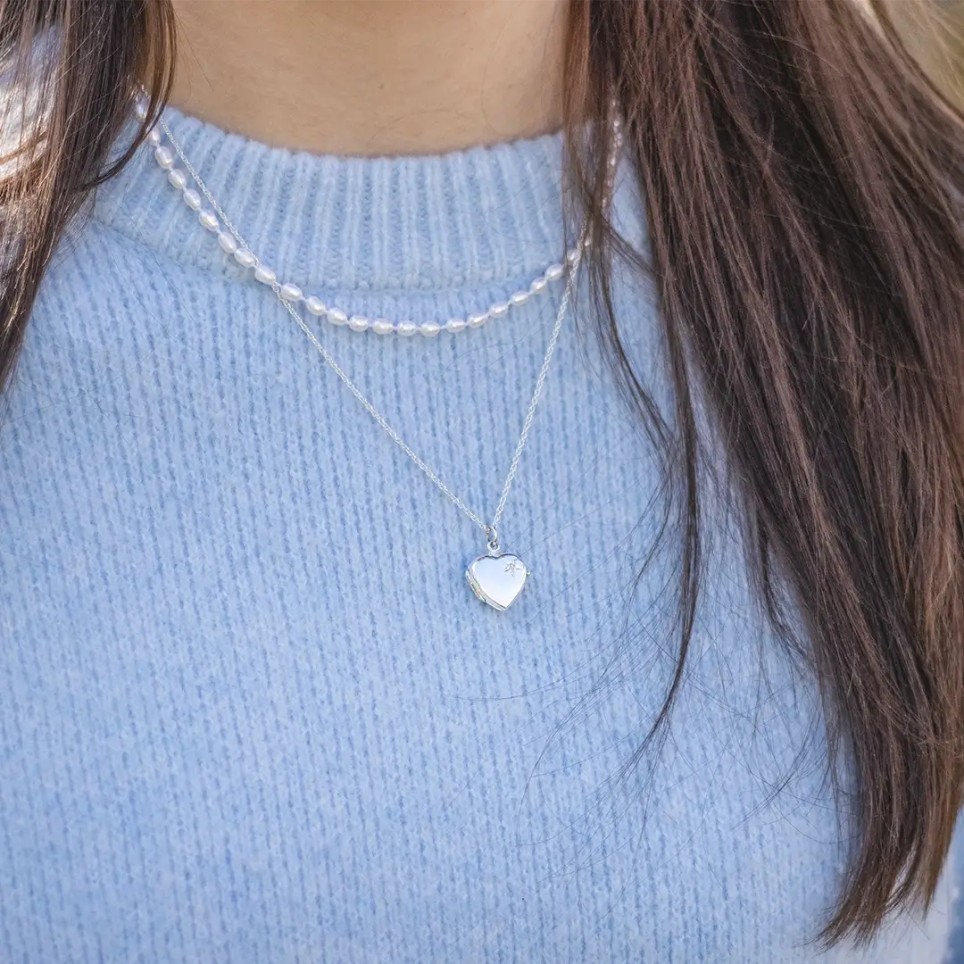 Close-up of a woman wearing a silver heart-shaped locket necklace layered with a pearl choker, styled over a light blue knit sweater.