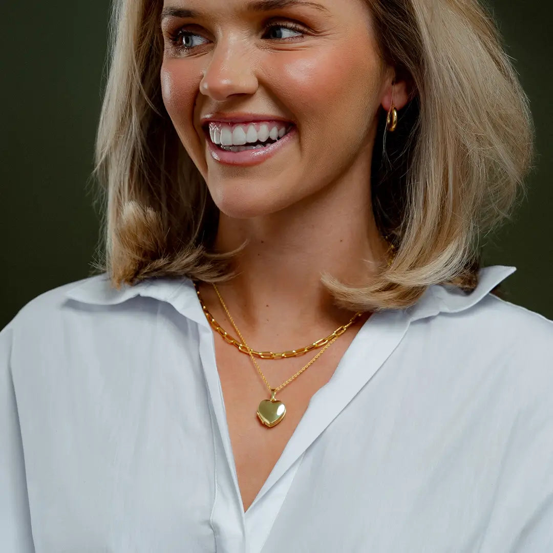 Model wearing a polished gold heart locket necklace layered with a gold chain, styled with a white blouse.