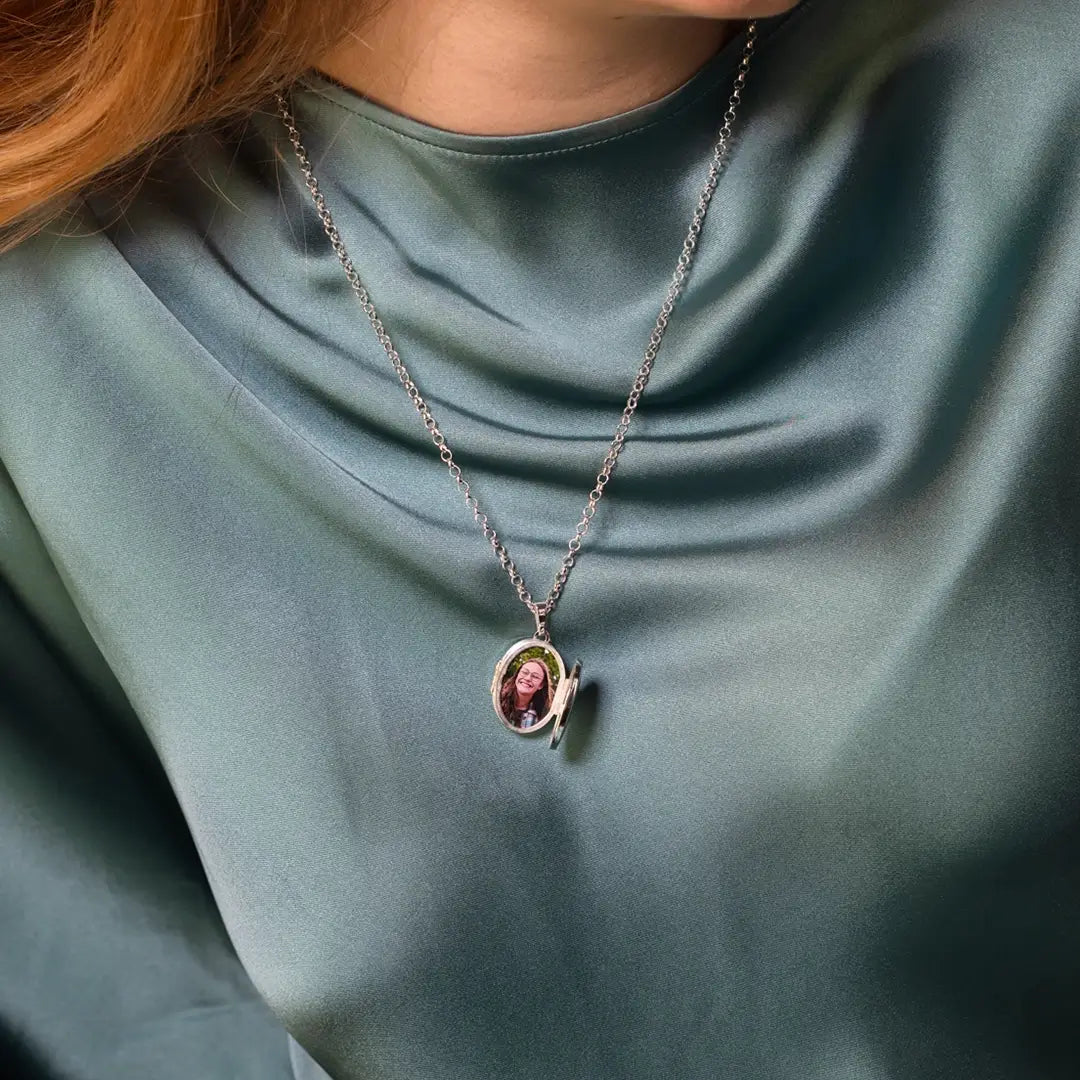 Sterling silver oval expanding photo locket worn on a chain, open to reveal a personalised portrait photograph inside, styled on a model wearing a satin green blouse.