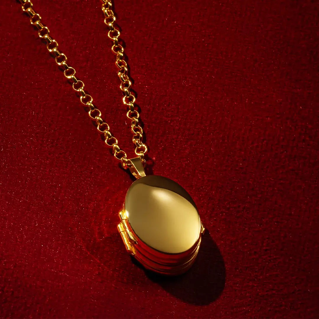 Gold oval locket necklace on a chain, photographed against a rich red textured background, highlighting the polished finish and classic hinged design.