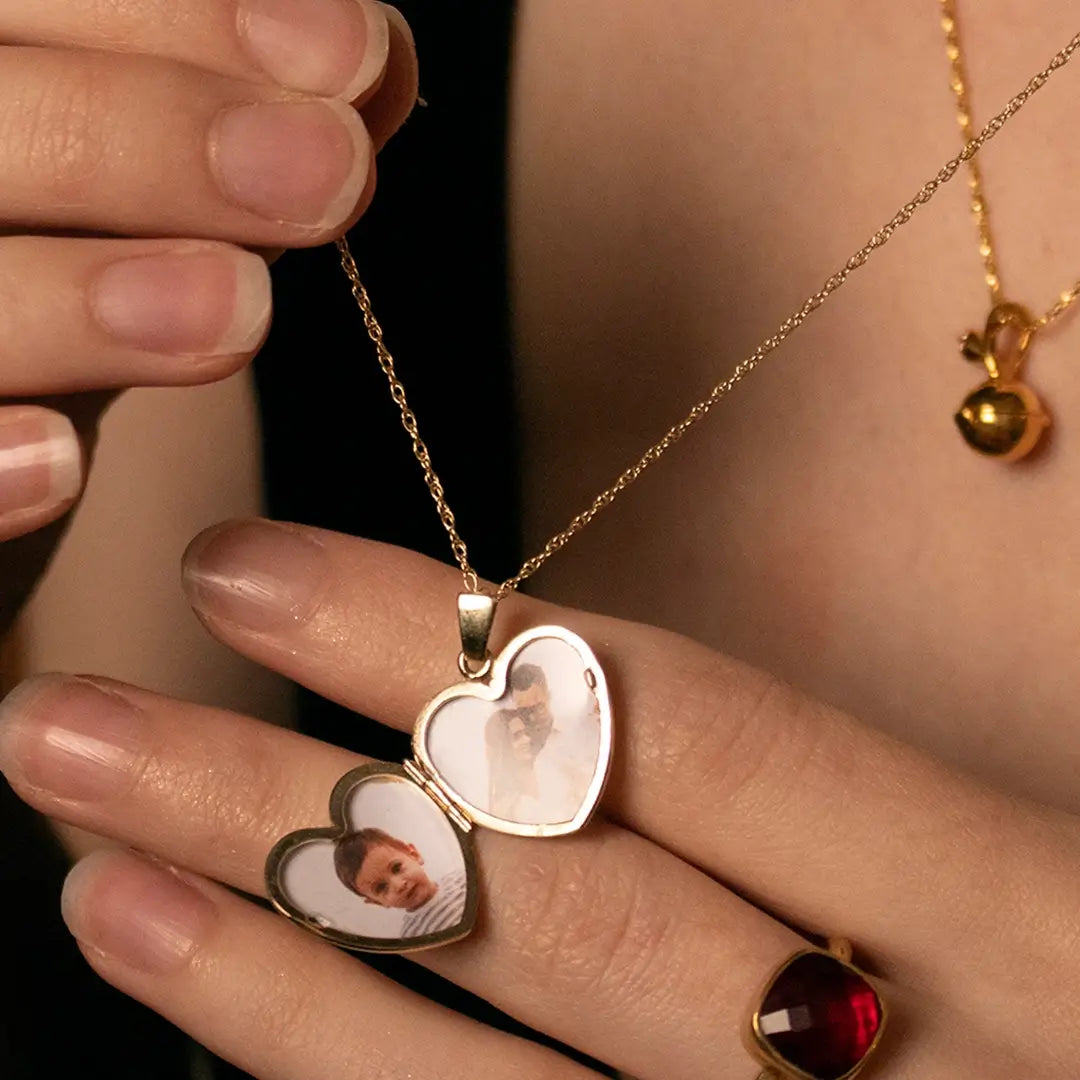 Gold heart-shaped photo locket necklace open to reveal two personalised photos, held in hand with delicate chain and gemstone ring