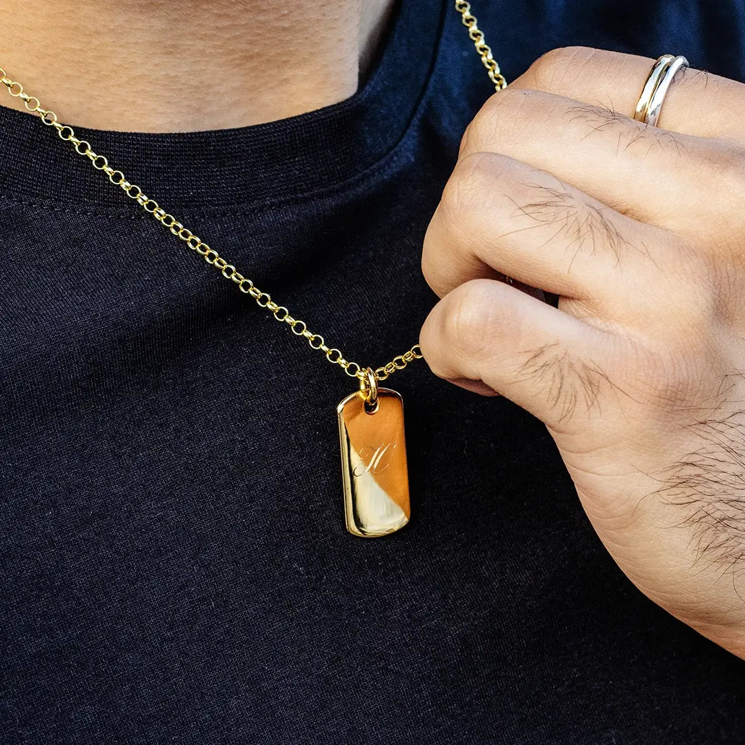 Close-up of a person holding a gold dog tag pendant necklace on a belcher chain, engraved with an initial, worn over a dark top