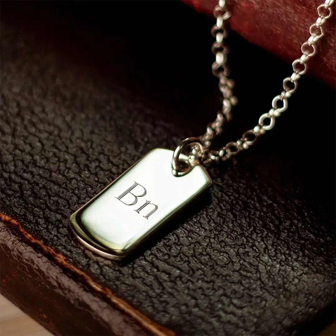 Engraved sterling silver dog tag necklace with initials on polished pendant, displayed on leather background with belcher chain.