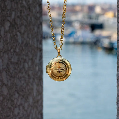 Gold compass locket necklace suspended outdoors between stone walls with soft blurred background