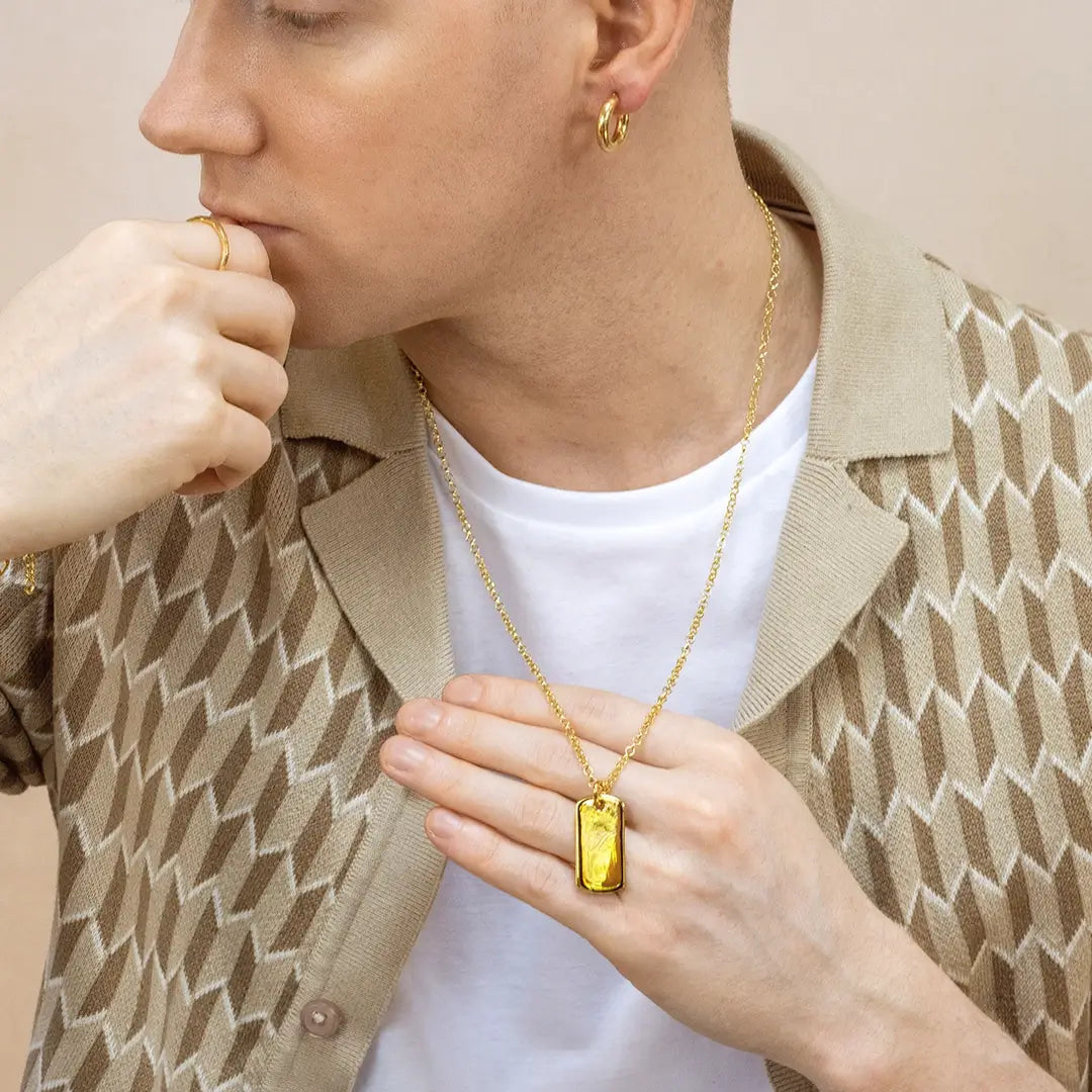 Person wearing a gold dog tag pendant necklace on a belcher chain over a white t-shirt and patterned jacket