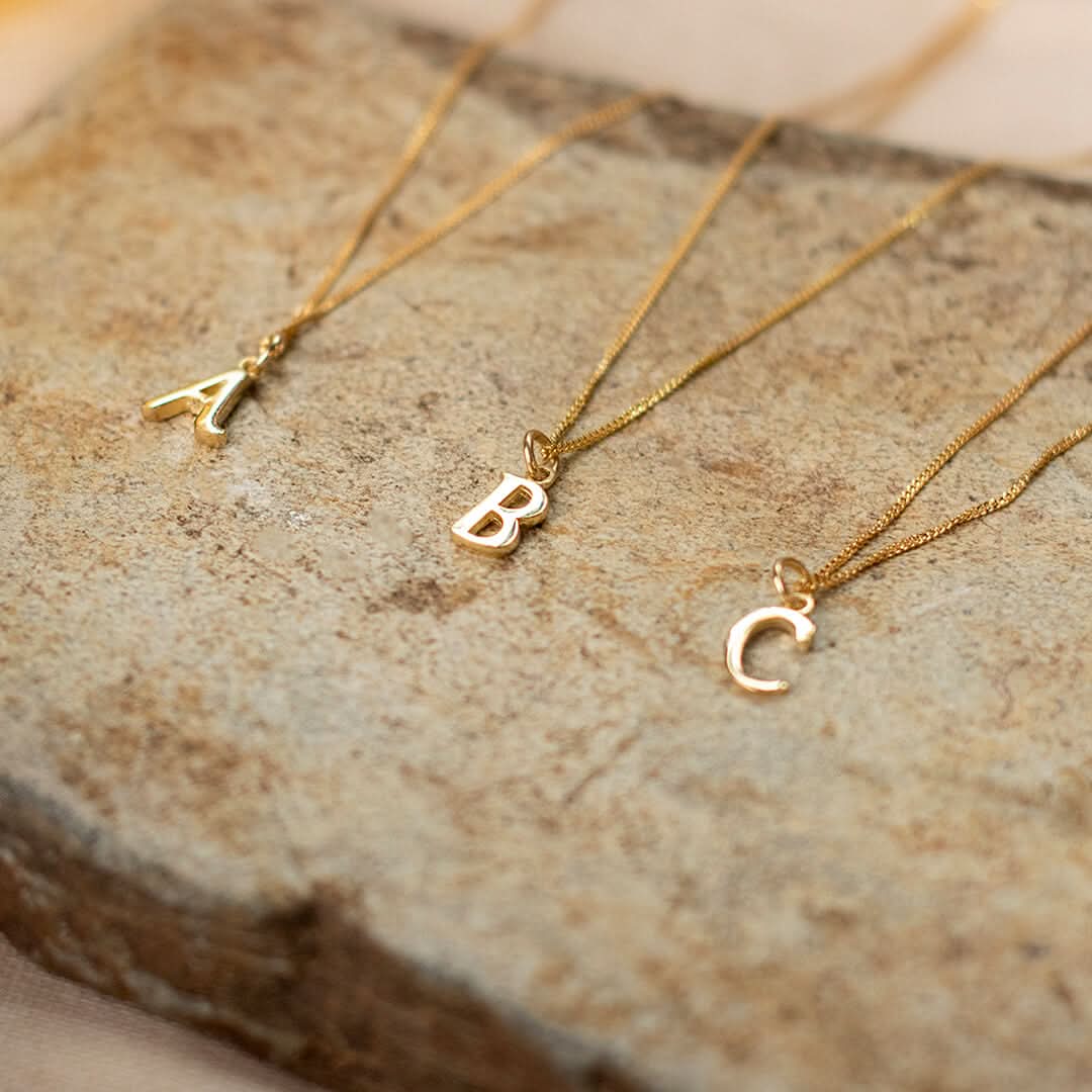 Three initial necklaces A, B and C,  in solid 9 carat gold on solid gold curb chains chains lying on a slab of textured stone.