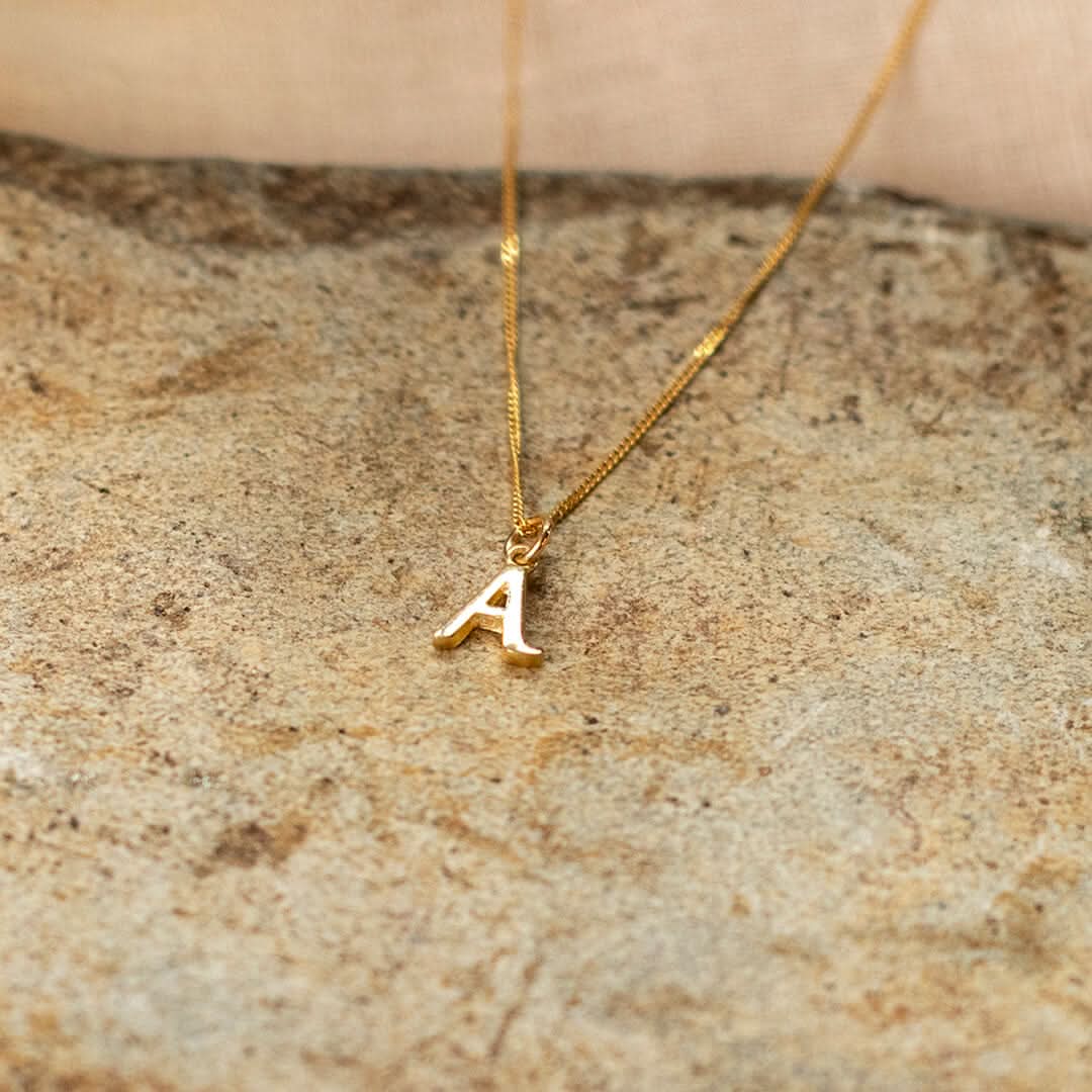  initial A necklace in solid gold on a curb chain, photographed lying on a sandstone slab. 