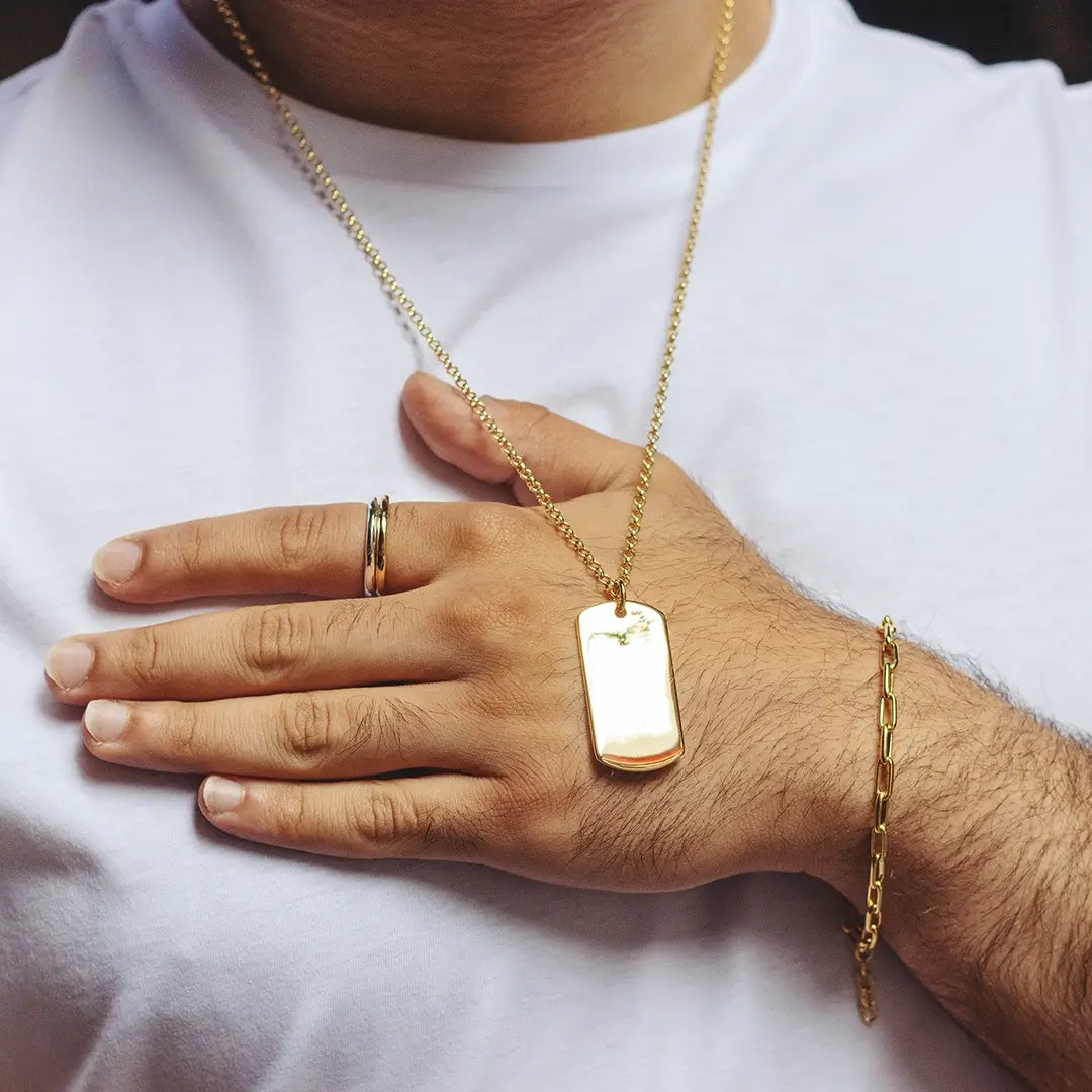 Close-up of a person wearing a gold dog tag pendant necklace, resting against their hand, styled with rings and a gold bracelet