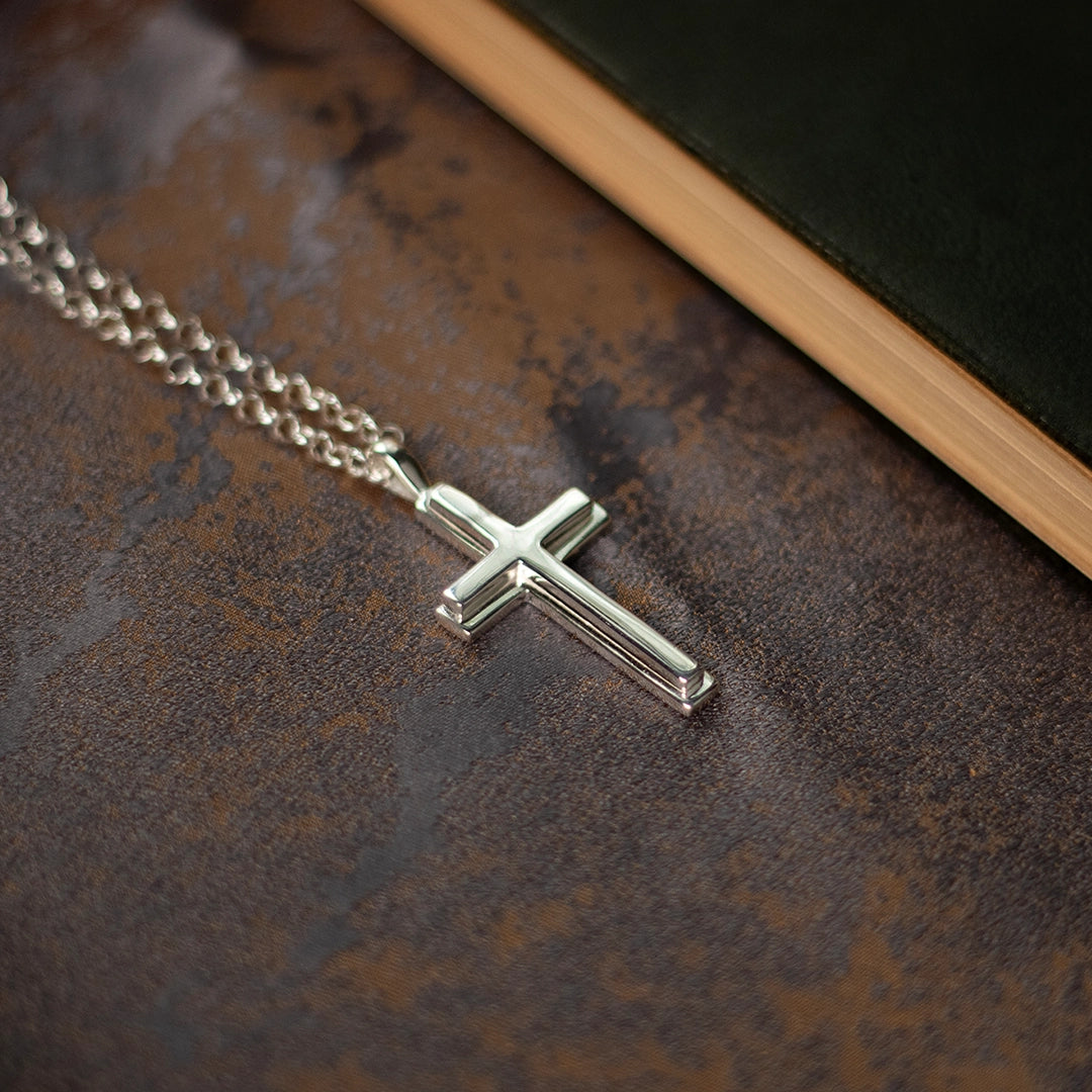 Sterling silver cross pendant necklace on chain, polished finish, styled on textured brown surface. By Lily Blanche.