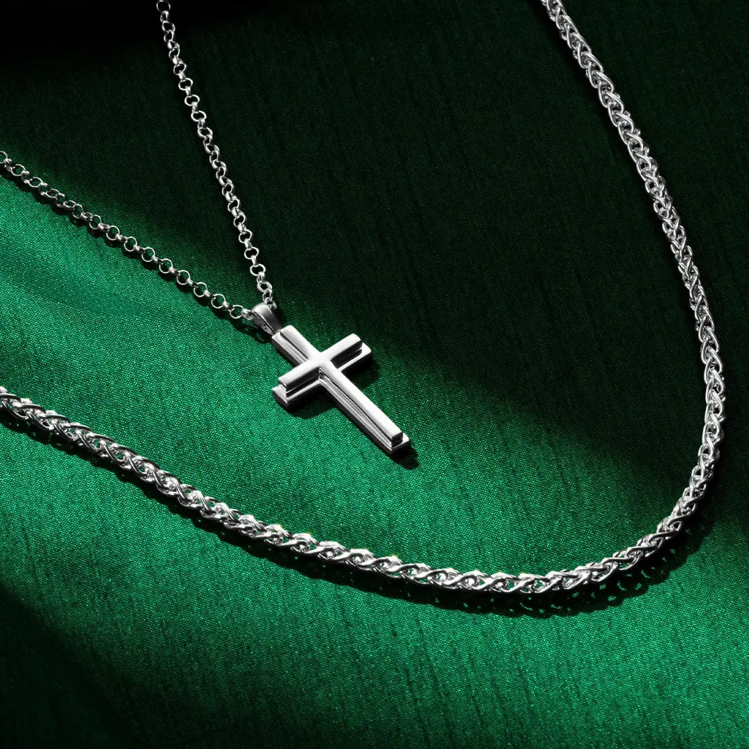 Silver cross pendant necklace layered beside a silver spiga chain on green satin fabric.