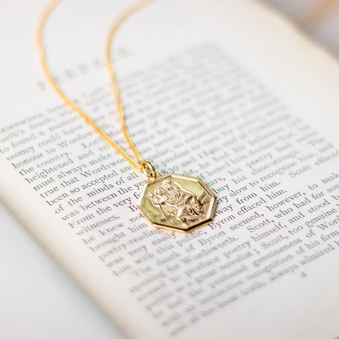 Gold St Christopher hexagon pendant necklace resting on open book pages, styled for meaningful gifting