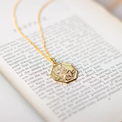 Gold St Christopher hexagon pendant necklace resting on open book pages, styled for meaningful gifting