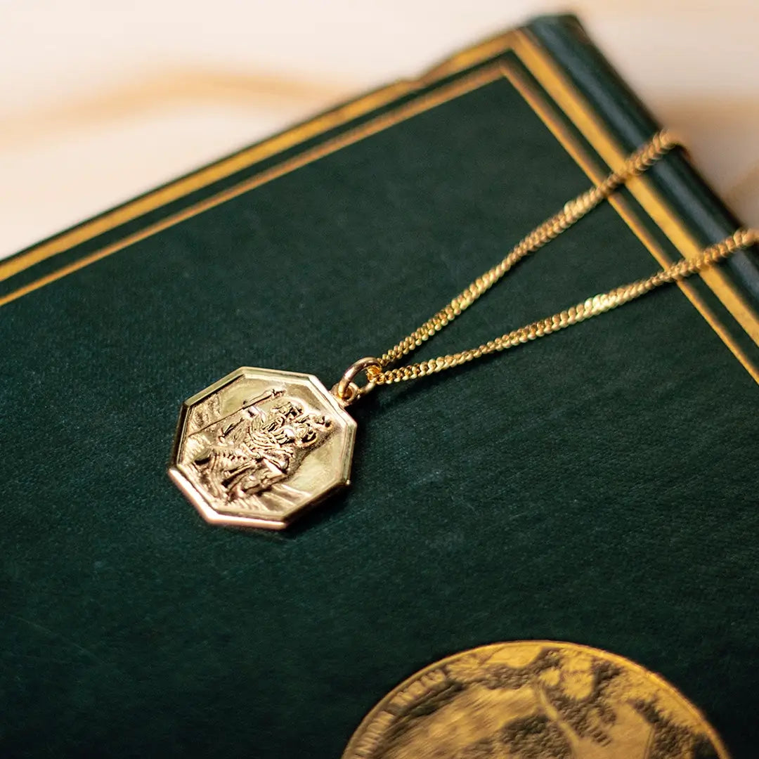 Gold hexagon St Christopher pendant necklace placed on vintage green book, highlighting heritage styling
