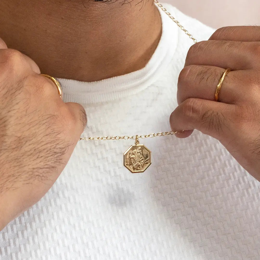 Men’s gold St Christopher hexagon pendant necklace held against white textured shirt, casual styling
