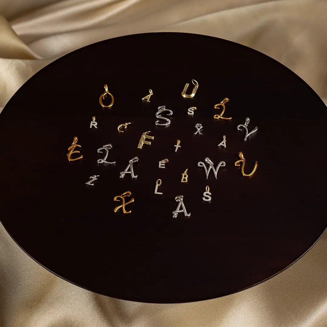 Luxury initial charm pendants in gold and silver arranged on a dark circular surface, featuring a variety of letter designs and fonts for personalised jewellery styling.
