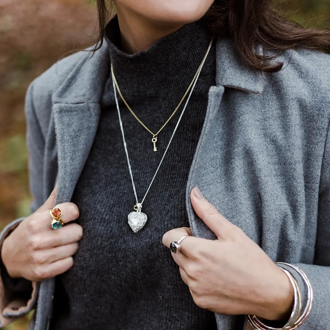 Close-up of a woman wearing layered necklaces, including a gold chain with a small key charm and a longer silver chain with a heart-shaped locket. She is dressed in a grey turtleneck and coat, accessorised with colourful gemstone rings and stacked bangles.