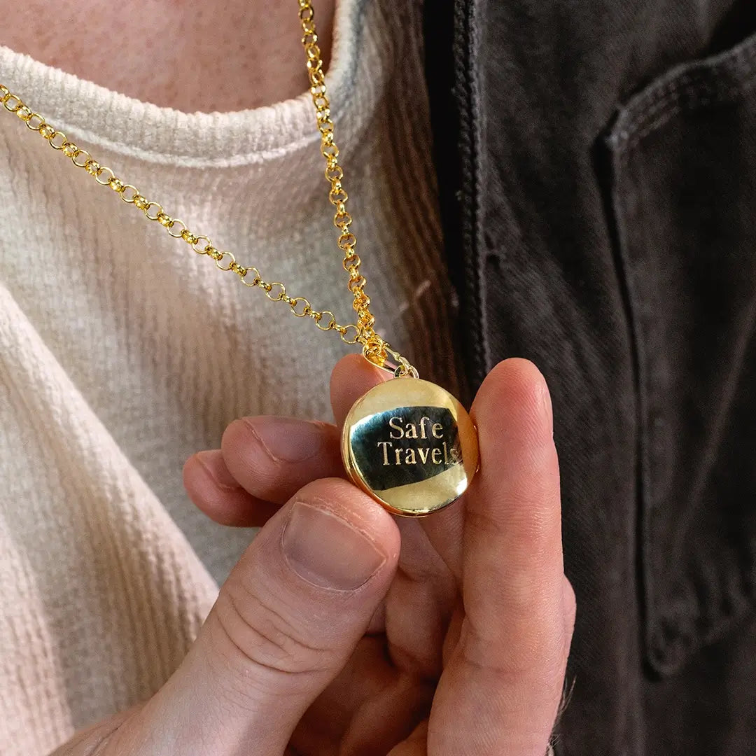 Close-up of a person holding a gold St Christopher locket showing engraved “Safe Travels” message on the back