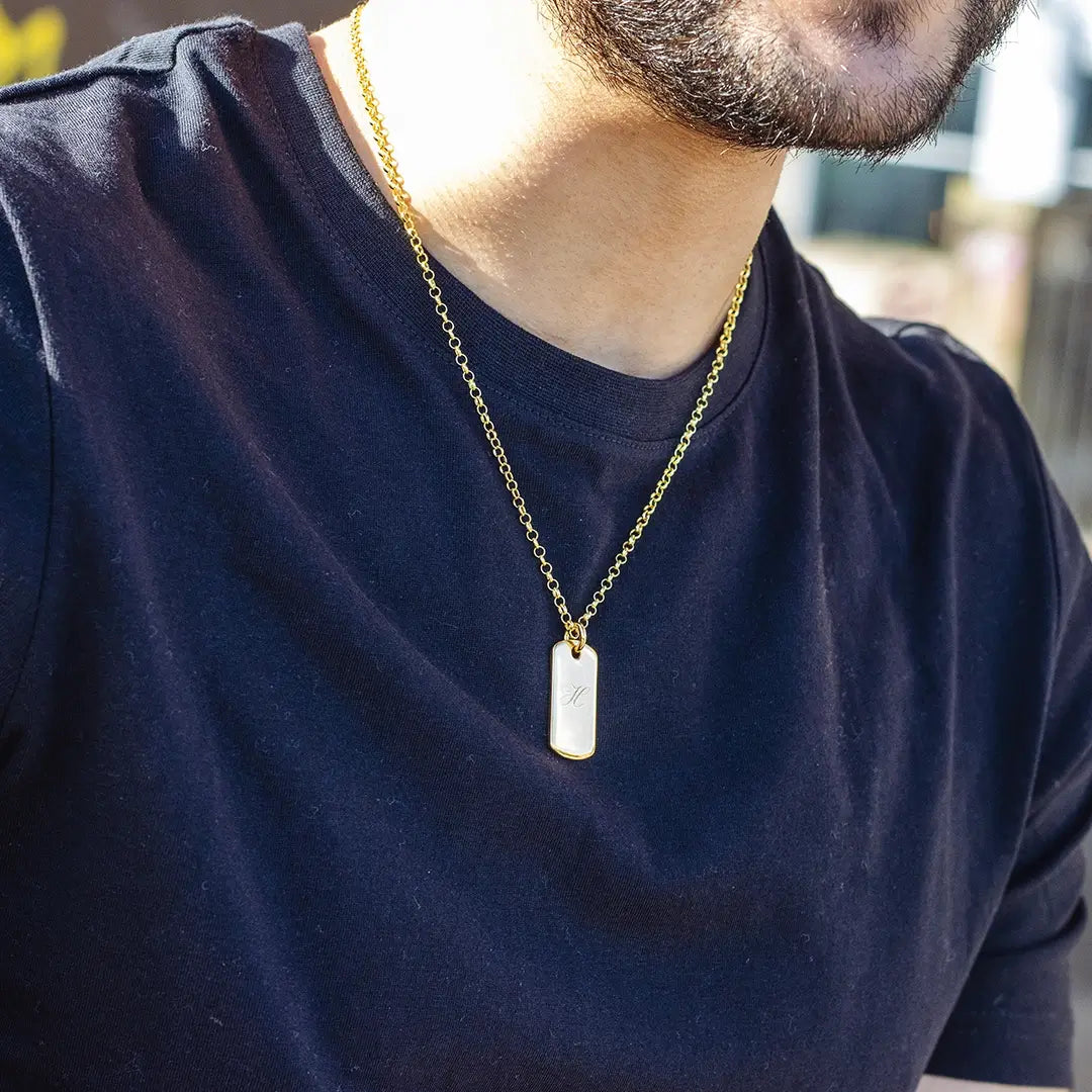 Close-up of a person wearing a gold dog tag pendant necklace engraved with an initial over a dark t-shirt