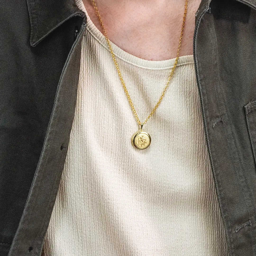 Close-up of a person wearing a gold St Christopher locket pendant necklace on a belcher chain over a textured cream top