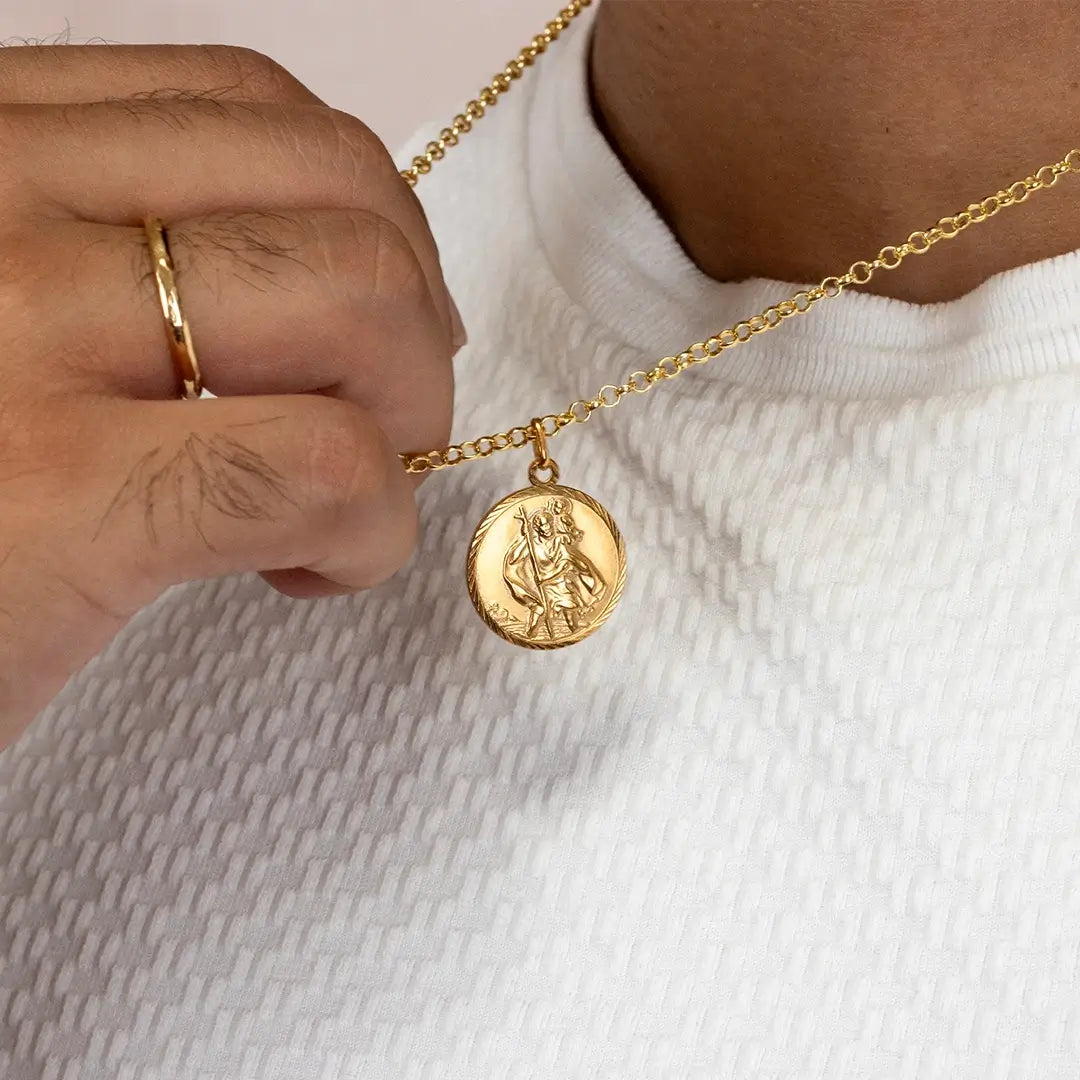 Close-up of gold St Christopher coin pendant necklace held against white shirt, highlighting engraved detail
