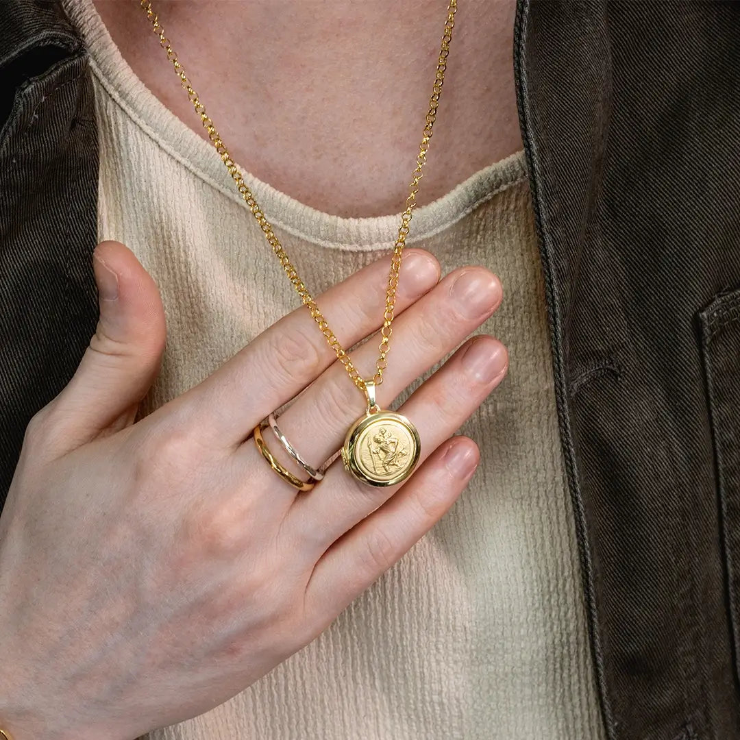 Close-up of a person holding a gold St Christopher locket pendant necklace on a belcher chain, worn with rings and a neutral outfit