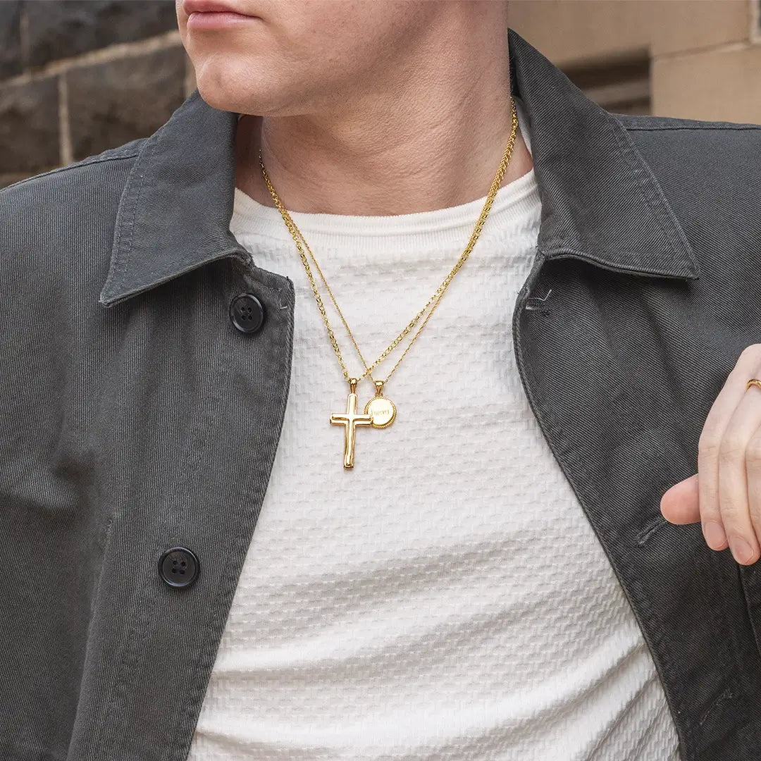 Mid-shot of a person wearing layered gold necklaces by Lily Blanche with a heavy gold cross pendant and engraved disc over a cream top and jacket