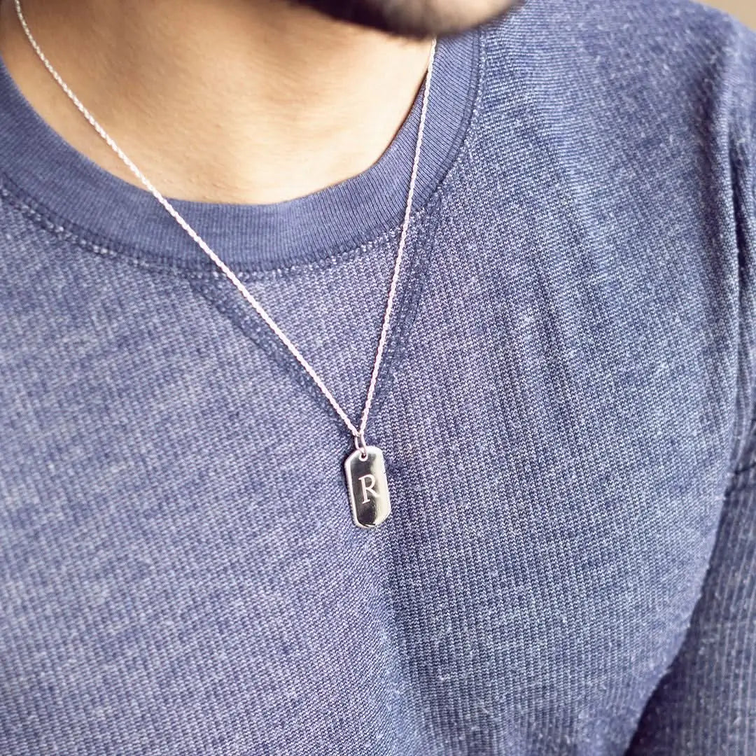Men’s sterling silver engraved dog tag necklace with initial pendant worn on belcher chain, styled on model in casual knitwear.