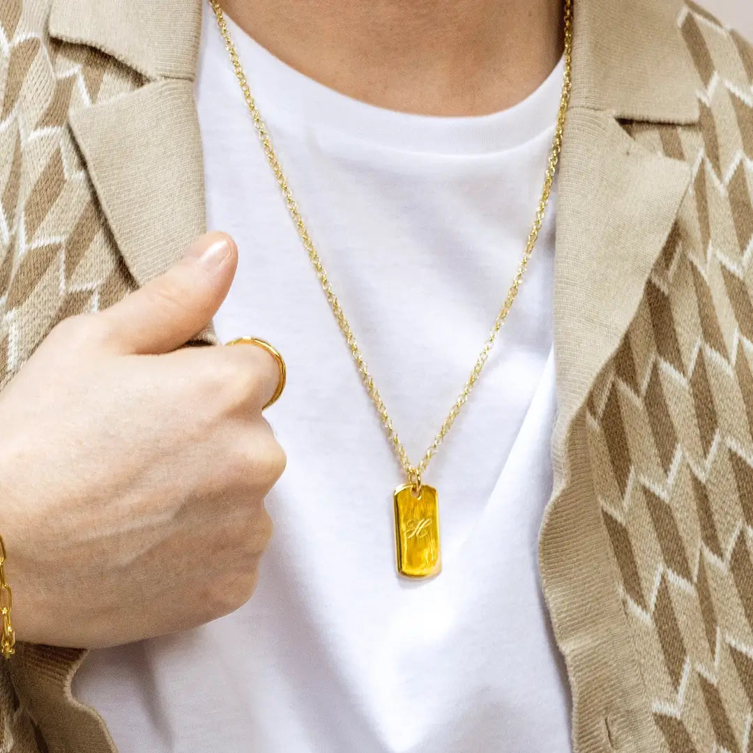 Person wearing a gold dog tag pendant necklace engraved with an initial over a white t-shirt and beige patterned jacket
