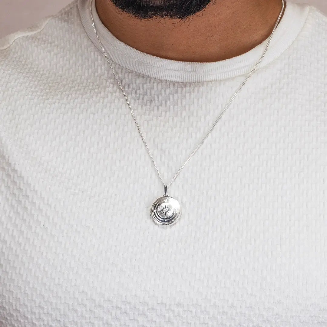Close-up of a person wearing a silver compass locket necklace on a fine chain over a textured white top