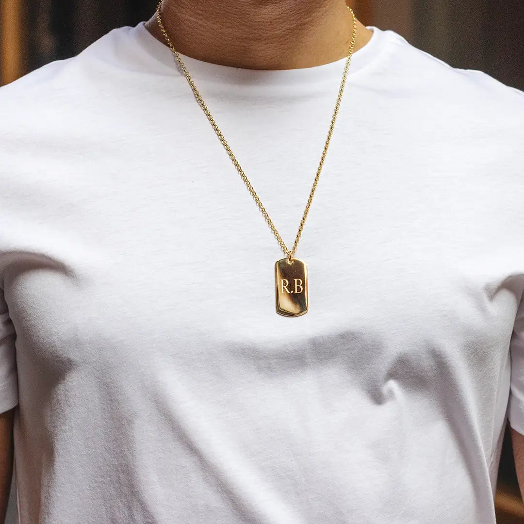 Person wearing a gold dog tag pendant necklace engraved with initials “R.B” on a fine chain over a white t-shirt