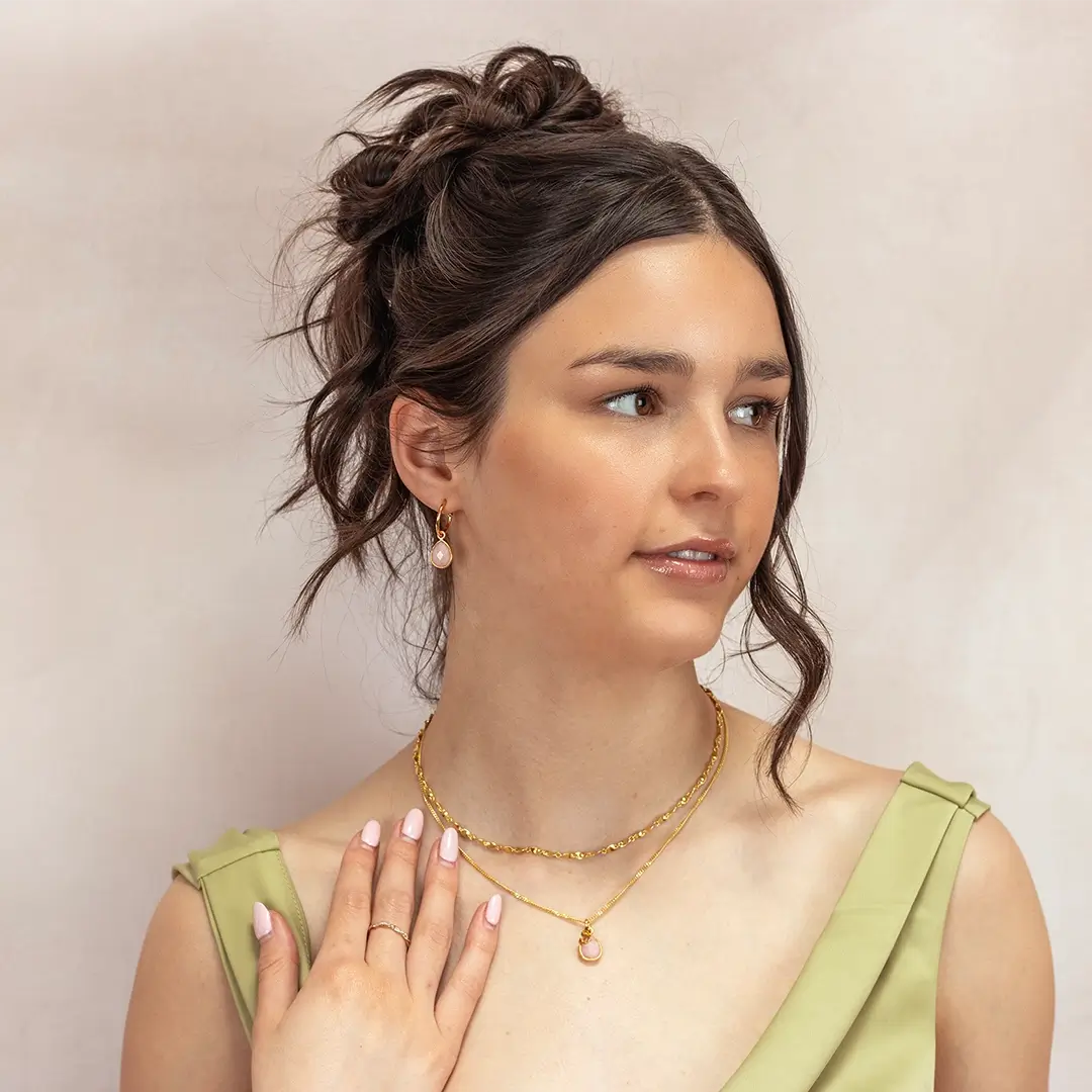 Woman wearing a matching gold jewellery set featuring a pink opal pendant necklace and dangling pink opal earrings. She touches her necklace with manicured nails and wears a green dress with pleated straps.