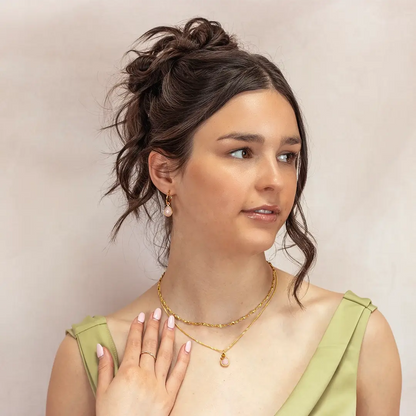 Woman wearing a matching gold jewellery set featuring a pink opal pendant necklace and dangling pink opal earrings. She touches her necklace with manicured nails and wears a green dress with pleated straps.