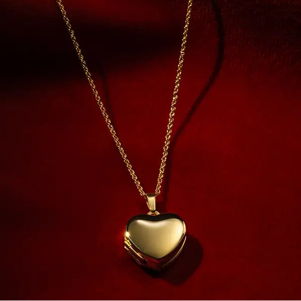 Polished gold heart locket necklace on a fine gold chain displayed on luxurious red fabric background.