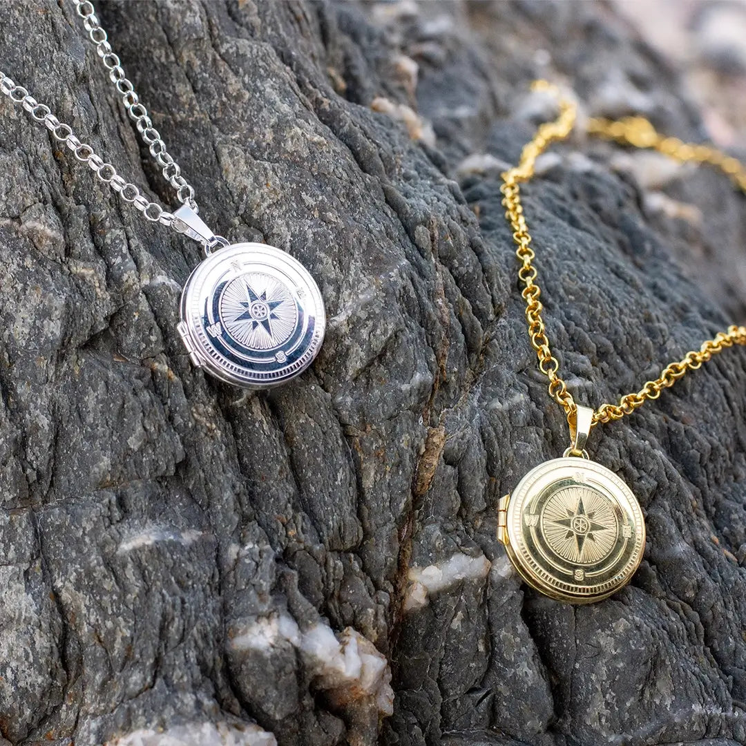 Silver and gold compass locket necklaces displayed on textured rock, highlighting contrast between metal finishes
