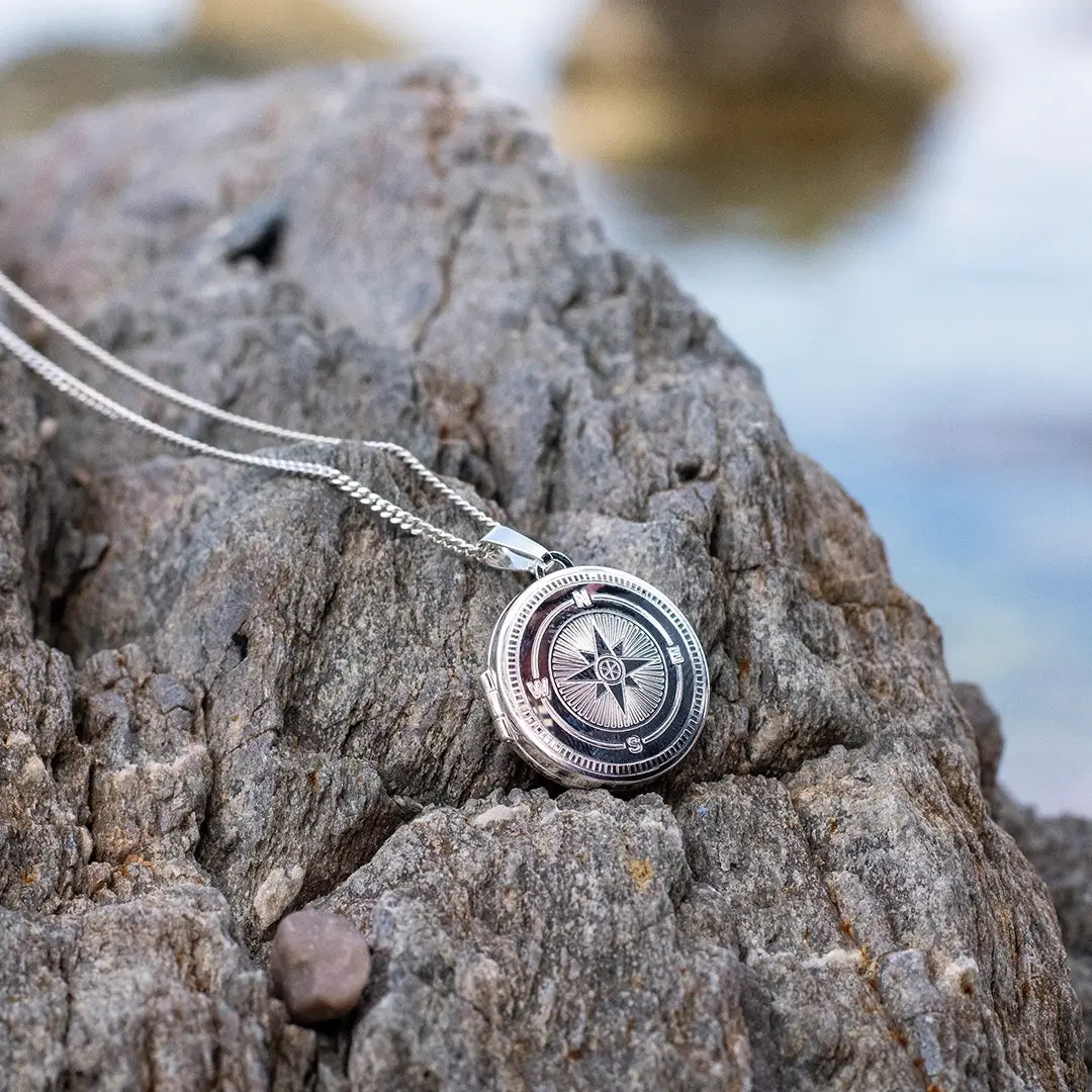 Close-up of a silver compass locket necklace resting on natural rock surface with water in the background