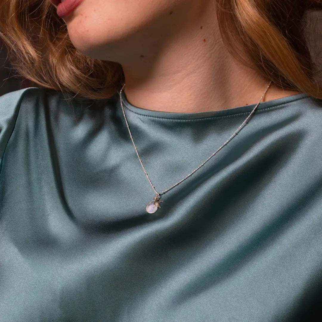 Woman wearing a silver chain necklace with a teardrop-shaped moonstone pendant, shown against a teal satin blouse