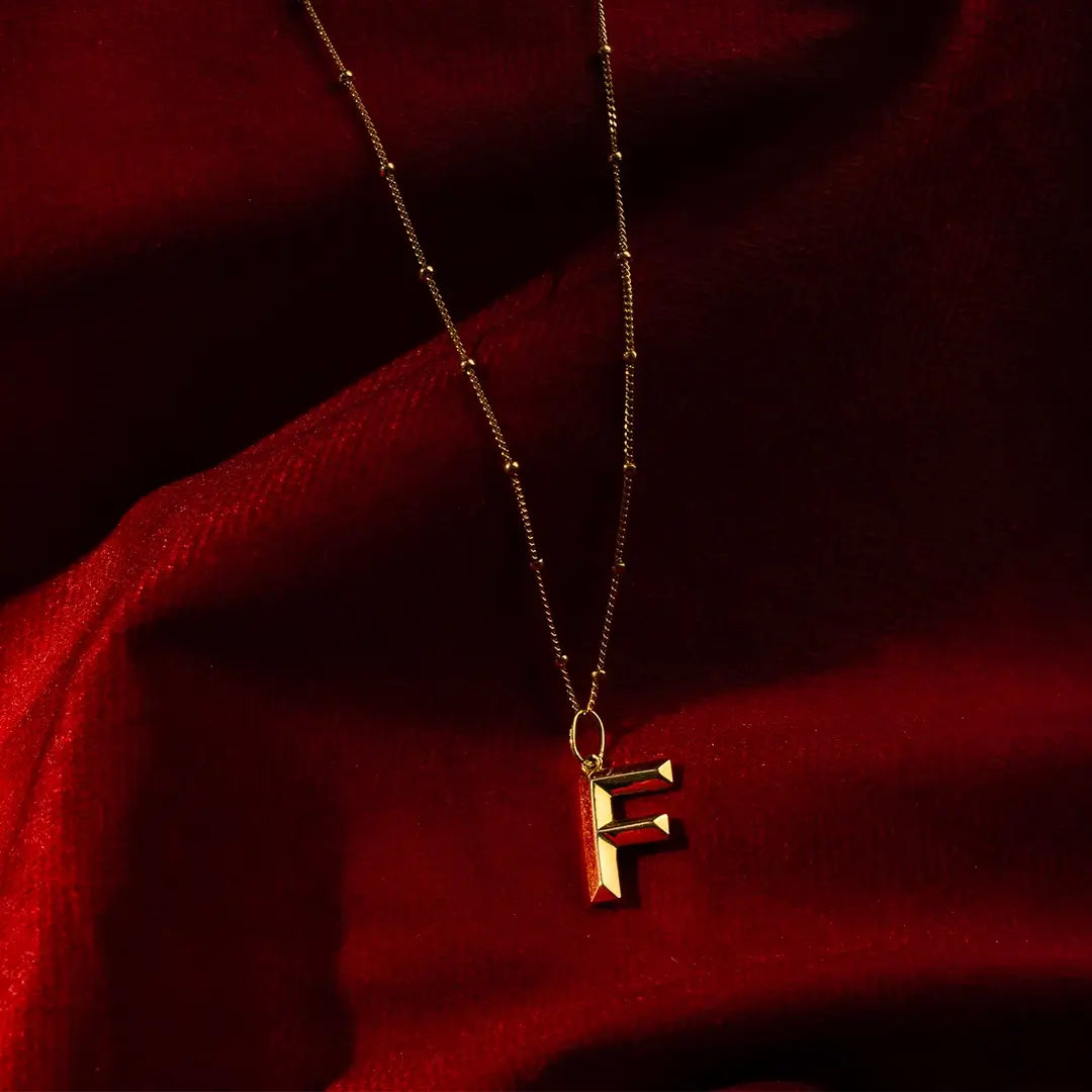 Solid 9ct gold initial necklace with a polished letter F pendant suspended from a delicate gold satellite chain, styled against rich red velvet fabric to highlight the warm gold finish and elegant design.
