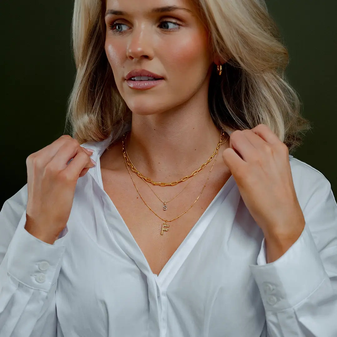 Model wearing a white blouse and layered gold necklaces, featuring a solid 9ct gold initial F pendant on a fine satellite chain. Styled with a chunky paperclip chain and a diamond initial necklace against a dark green background.