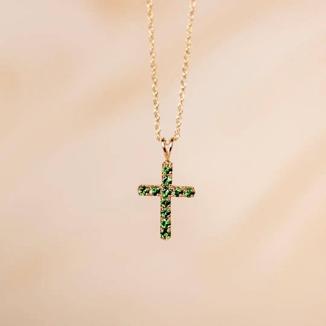 Gold cross pendant necklace featuring vivid green tsavorite gemstones on a fine chain against a soft