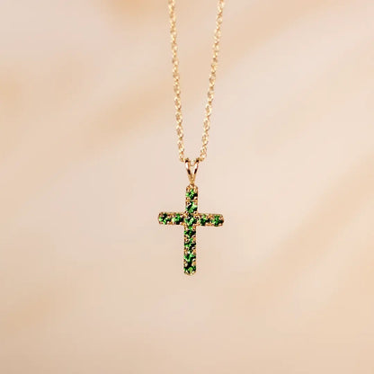 Gold cross pendant necklace featuring vivid green tsavorite gemstones on a fine chain against a soft