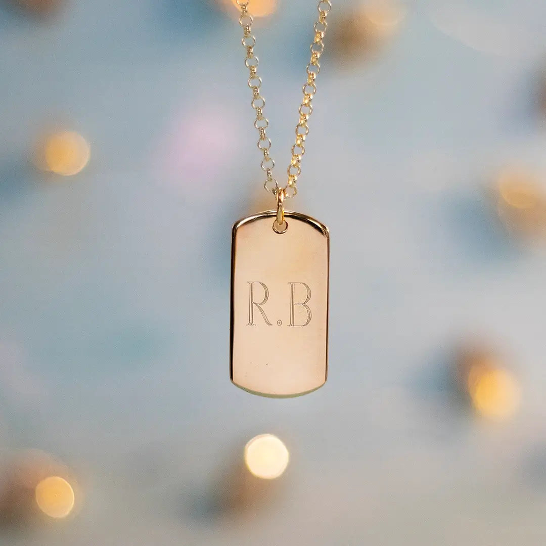 Gold dog tag pendant necklace engraved with initials “R.B” on a fine chain against a soft blurred background