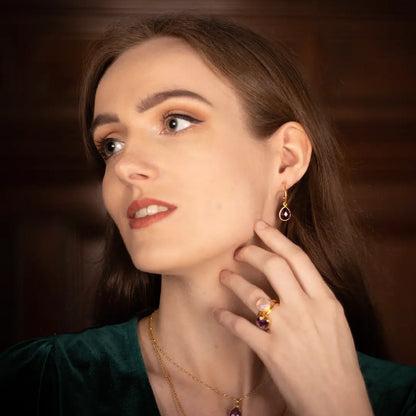 Woman wearing gold vermeil drop earrings with pear-cut amethyst gemstone, matching amethyst ring and necklace, styled with an emerald green velvet top.
