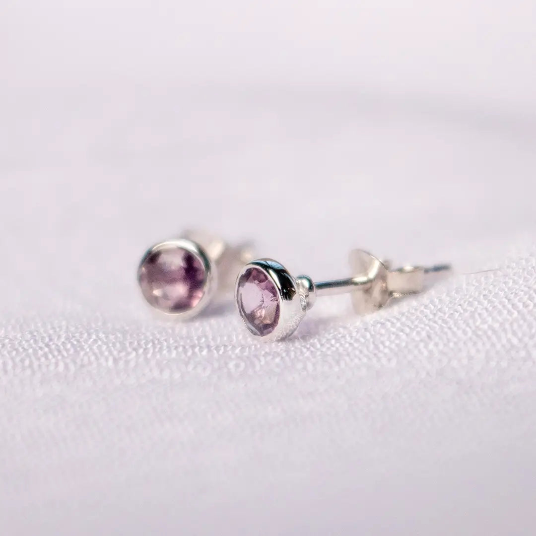 Close-up of sterling silver stud earrings with round-cut amethyst gemstones in a polished bezel setting on a soft white fabric background.