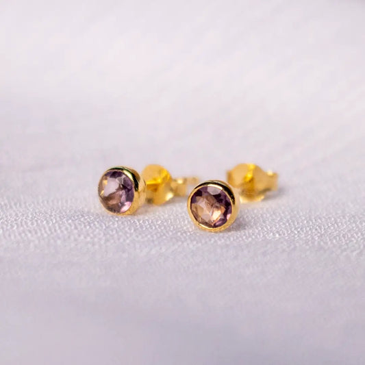 Close-up of gold vermeil stud earrings with round-cut amethyst gemstones, set in a polished bezel on a soft neutral background.