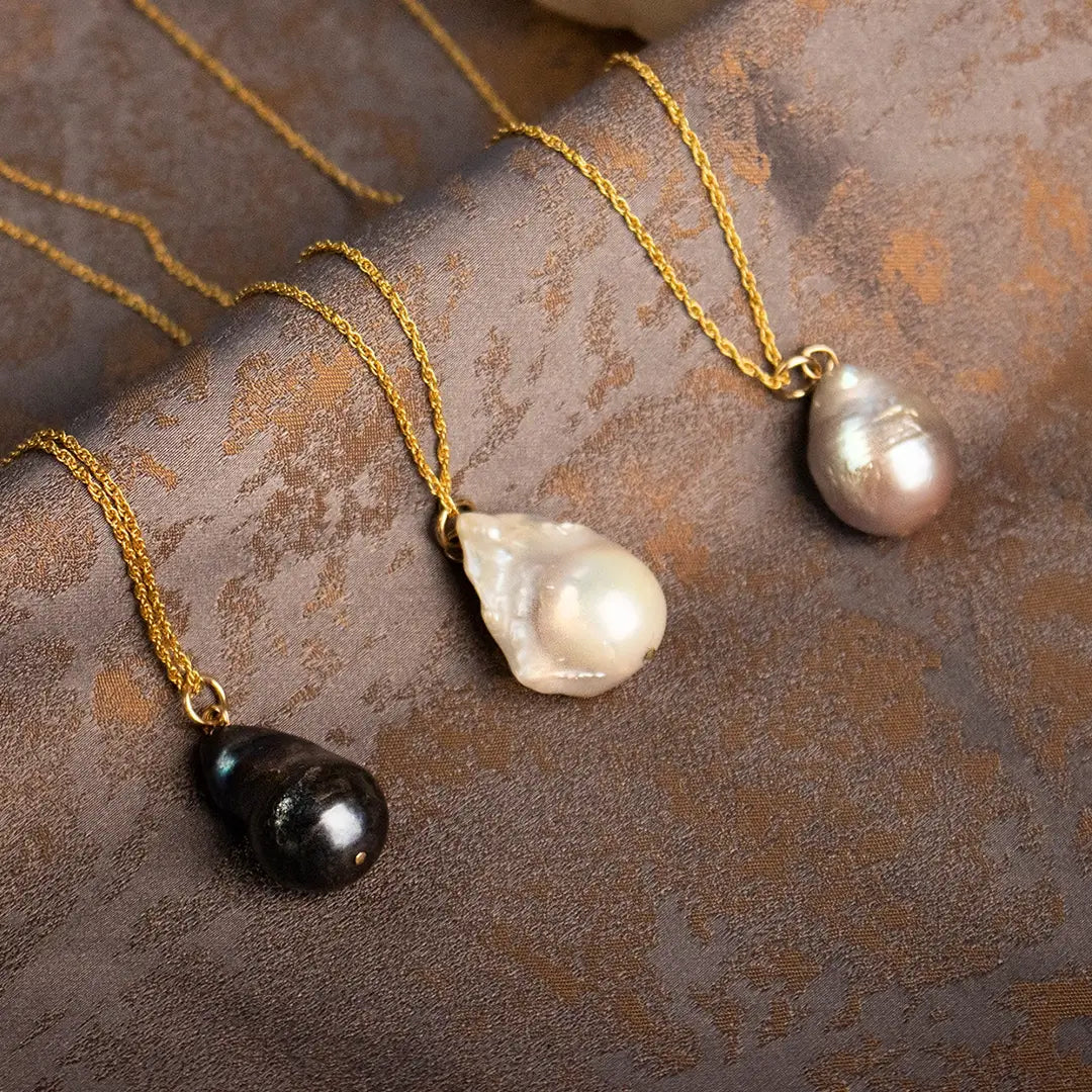 Three baroque pearl pendant necklaces on gold chains featuring black, ivory and blush pearls, displayed on textured neutral fabric.