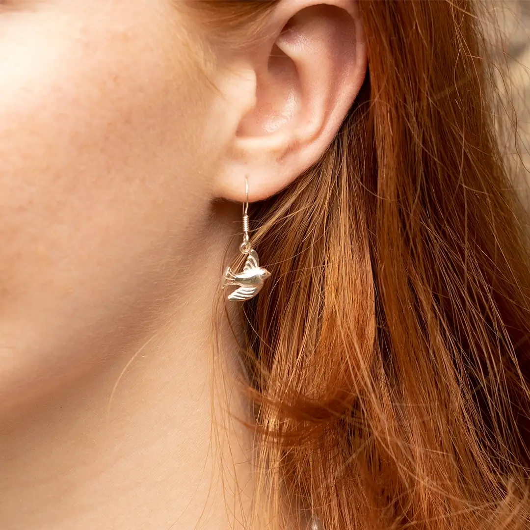 Close-up of a sterling silver bird drop earring worn on the ear, featuring a polished flying bird charm on a delicate hook fastening.